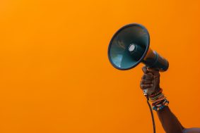 Black hand holding Megaphone on orange background. Isolated loudspeaker announcement and communication creative banner concept